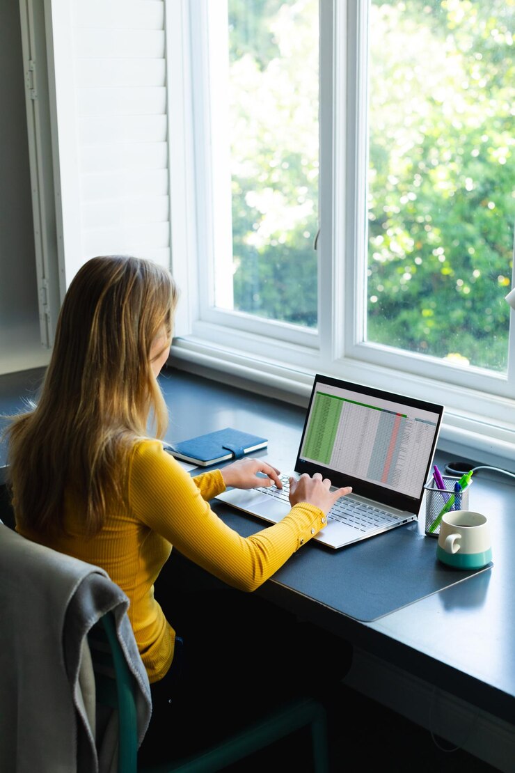 Foto mujer caucásica sentada en un escritorio con café trabajando desde casa y usando una computadora portátil en una habitación soleada