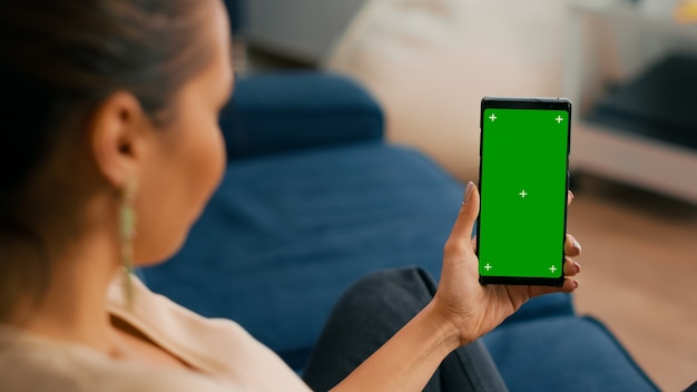 Mujer caucásica sentada en el sofá en la sala de estar mientras se socializa en la aplicación de Internet usando el teléfono con pantalla verde de maqueta chroma key display.Mujer navegando en red con gadget aislado