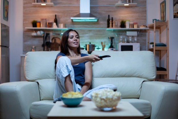 Mujer caucásica sentada en el sofá y viendo la televisión relajándose después del trabajo. Señora sola en casa divertida emocionada en pijama descansando con bocadillos y jugo sentado en un cómodo sofá en la sala de estar de espacio abierto.