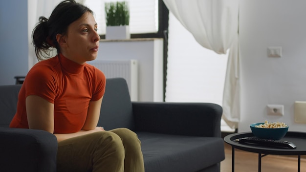 Mujer caucásica sentada sola en el sofá en la sala de estar comiendo palomitas de maíz mientras espera que su marido se siente. Persona joven en el sofá hablando con su pareja mientras toma un refrigerio de la mesa de café