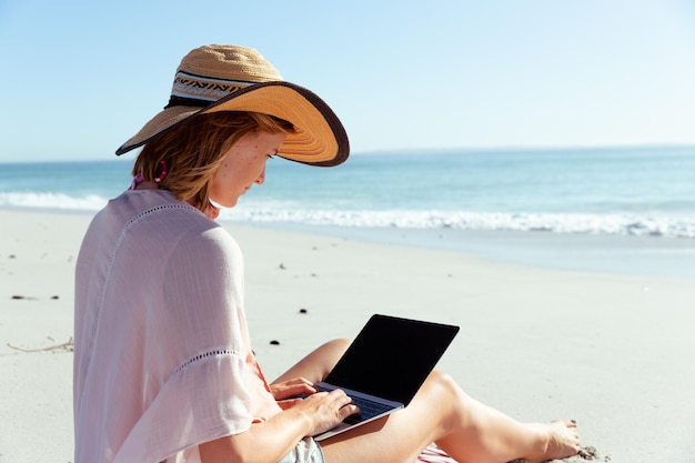 Una mujer caucásica con sombrero, disfrutando del tiempo en la playa en un día soleado, usando una computadora portátil, con el mar de fondo