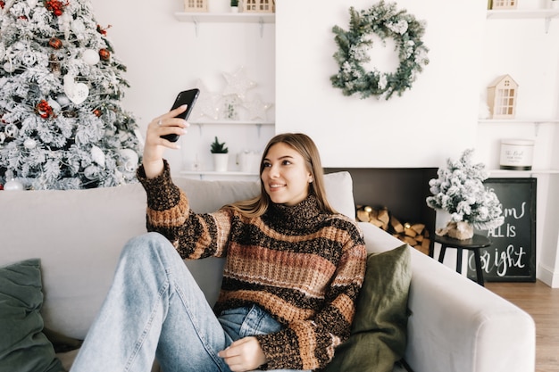 Mujer caucásica sonriente joven que hace selfie usando el teléfono móvil cerca del árbol de Navidad en casa.