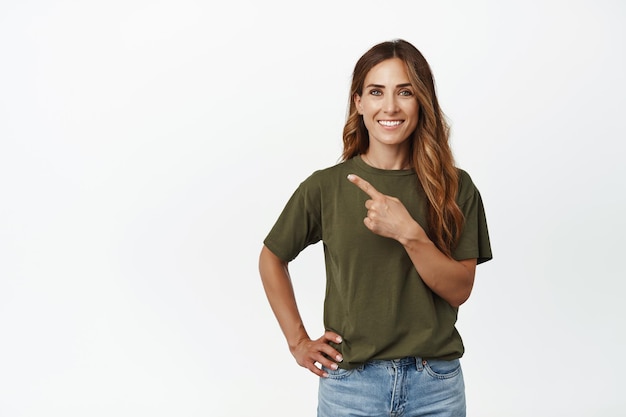 Mujer caucásica sonriente que se ve feliz, señalando con el dedo en la esquina superior izquierda, mostrando publicidad, recomienda venta promocional, parada en camiseta y jeans contra fondo blanco