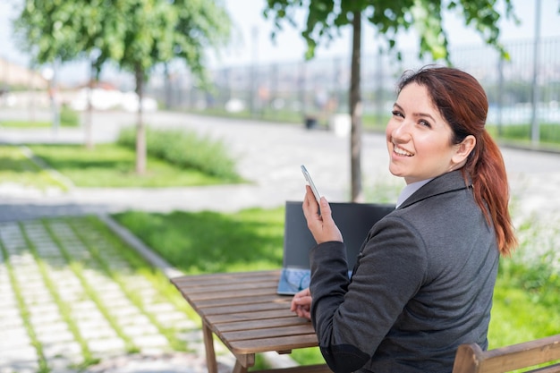 Una mujer caucásica sonriente se sienta en una mesa de madera en una terraza de verano y usa un teléfono inteligente