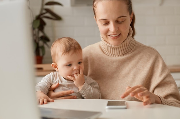 Mujer caucásica sonriente con suéter beige sentada en la cocina y trabajando en línea en un teléfono inteligente o revisando sus redes sociales pasando tiempo con su hija