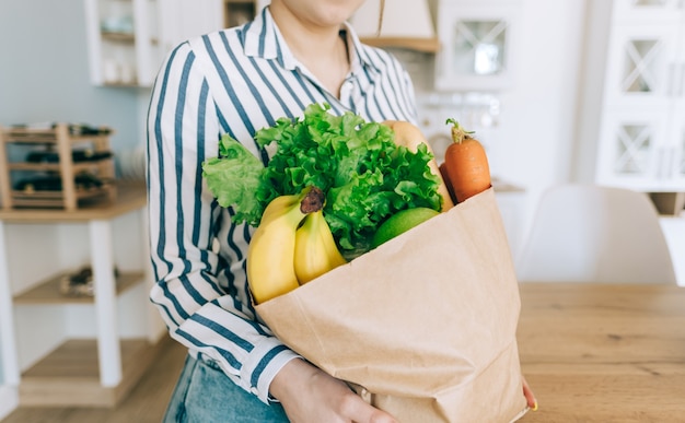 Mujer caucásica sostenga la bolsa de compras ecológica con verduras frescas y baguette en la cocina moderna en casa.