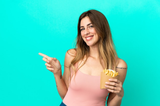 Mujer caucásica sosteniendo papas fritas aisladas sobre fondo azul apuntando con el dedo hacia el lado