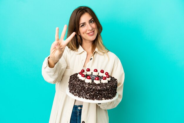 Mujer caucásica sosteniendo pastel de cumpleaños aislado sobre fondo azul feliz y contando tres con los dedos