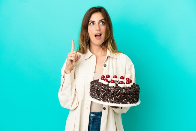 Foto mujer caucásica sosteniendo pastel de cumpleaños aislado sobre fondo azul pensando en una idea apuntando con el dedo hacia arriba