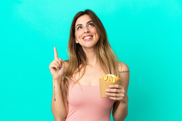Mujer caucásica sosteniendo patatas fritas aisladas sobre fondo azul apuntando hacia una gran idea