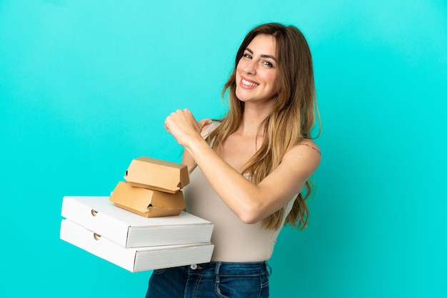 Mujer caucásica sosteniendo pizzas y hamburguesas aislado sobre fondo azul orgulloso y satisfecho de sí mismo