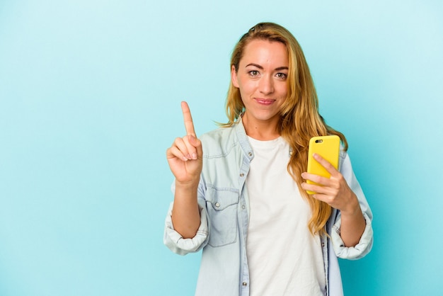 Mujer caucásica sosteniendo teléfono móvil aislado sobre fondo azul mostrando el número uno con el dedo.
