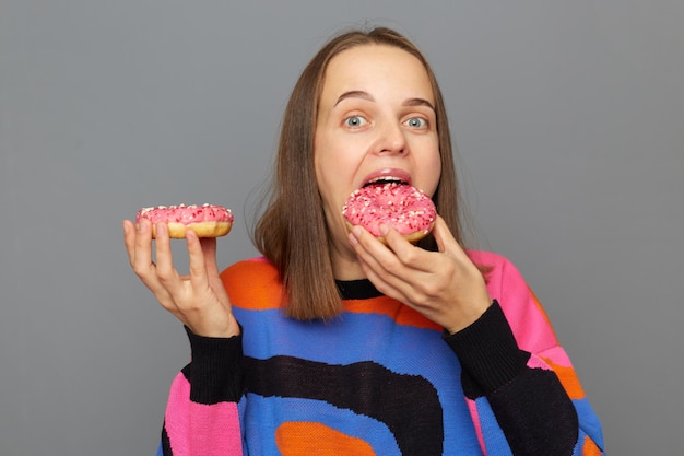 Mujer caucásica con suéter posando aislada sobre fondo gris muerde un delicioso donut con chispitas brillantes tiene una nutrición poco saludable no puede imaginar la vida sin postres dulces no sigue la dieta