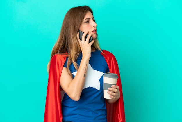 Mujer caucásica de superhéroe aislada en la pared azul con café para llevar y un móvil