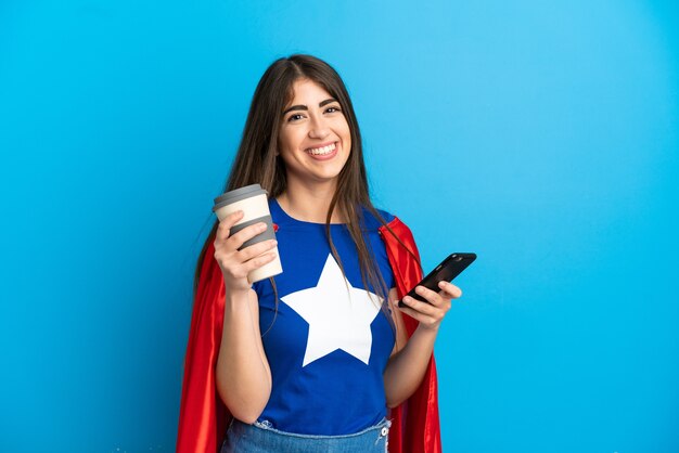 Mujer caucásica de superhéroe aislada sobre fondo azul con café para llevar y un móvil