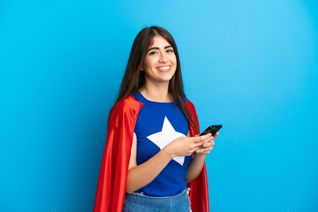 Mujer caucásica de superhéroe aislada sobre fondo azul enviando un mensaje con el móvil