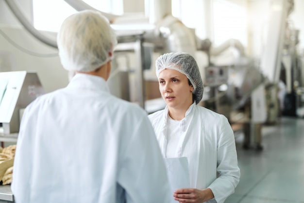 Mujer caucásica en uniforme estéril con papeleo y hablando con el empleado mientras está de pie en la planta de alimentos. En máquinas de fondo.