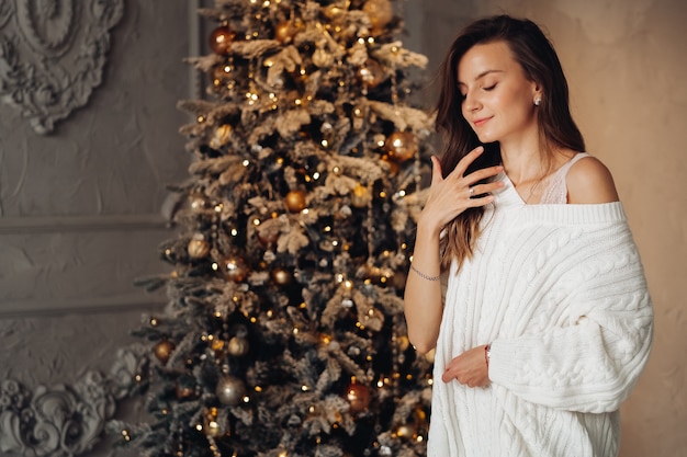 Mujer caucásica en vestido y medias posa cerca del árbol de Navidad con muchos regalos y sonrisas