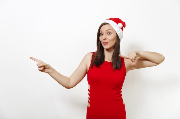 Foto mujer caucásica con vestido rojo, sombrero de navidad, apuntando a un lado con los dedos índices en ambas manos aisladas sobre fondo blanco. santa niña. concepto de vacaciones de año nuevo 2018. copie el espacio para publicidad.