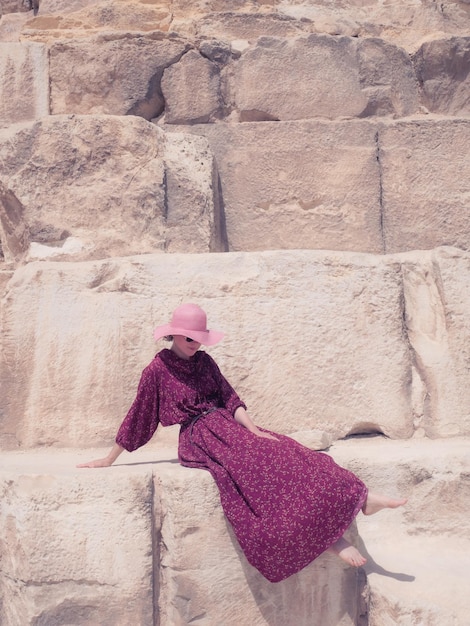 Una mujer caucásica con un vestido rojo y un sombrero se sienta maravillosamente en la pirámide de Khafre en Giza.