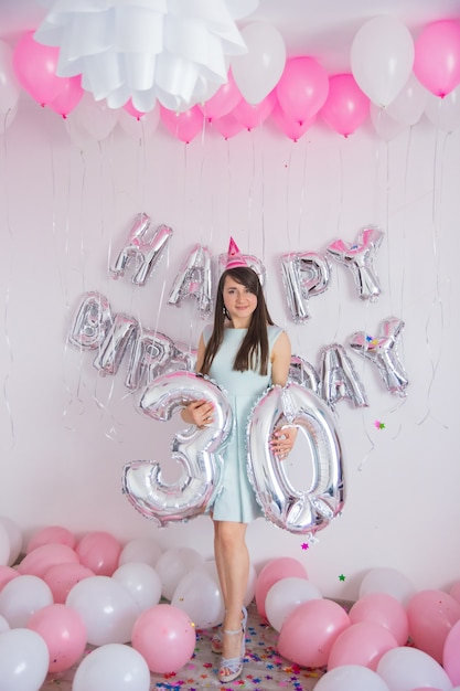 La mujer celebra su 30 cumpleaños. Decoraciones de cumpleaños con globos de color blanco y rosa y confeti para fiesta sobre un fondo de pared blanca. Globo concepto 30 años. Número treinta de plata