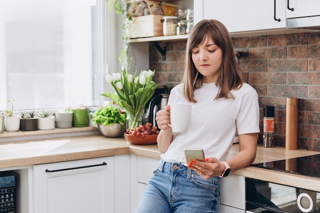 Mujer de cerca en camiseta blanca tomando café o té y usando el teléfono en la cocina de casa. Desayuno temprano en la mañana, tiempo de descanso, descanso del trabajo.