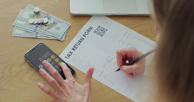 Una mujer de cerca llena un documento en papel y cuenta en la pantalla del teléfono Una cliente joven está escribiendo un formulario de declaración de impuestos y mirando una computadora portátil en la mesa en la habitación de su casa