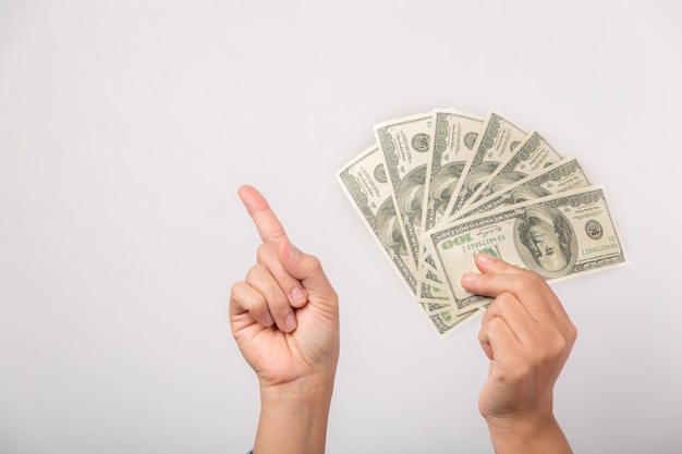 Una mujer de cerca sosteniendo dólares feliz señalando con la mano y el dedo hacia el lado con éxito mostrando dinero y jactándose de ganar la lotería con grandes ganancias en un estudio interior aislado en fondo gris