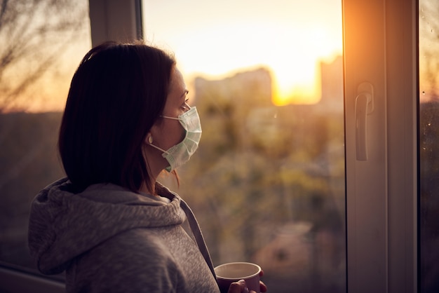 Mujer cerca de la ventana al atardecer en aislamiento en casa por brote de virus. Concepto de quedarse en casa