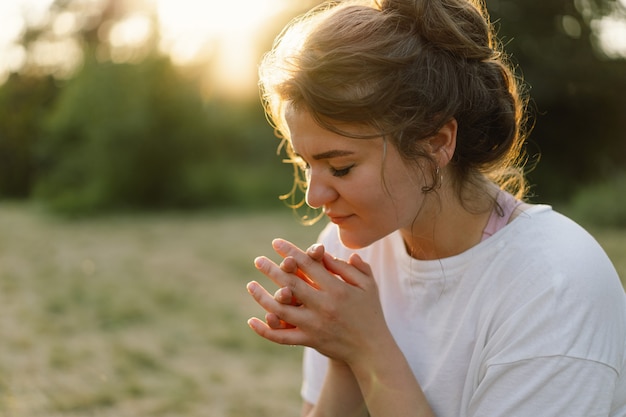 La mujer cerró los ojos rezando en un campo durante la hermosa puesta de sol manos dobladas en concepto de oración por ...
