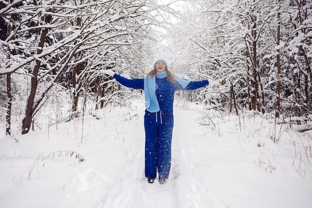 Mujer en un chándal azul guantes blancos y bufanda para arrojar nieve en invierno en un bosque cubierto de nieve