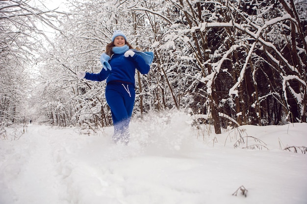 Mujer en un chándal azul, mitones blancos y bufanda se encuentra en invierno en un bosque cubierto de nieve