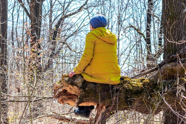 Mujer en una chaqueta amarilla sentada en el bosque en un árbol caído, relajarse mientras camina en el bosque