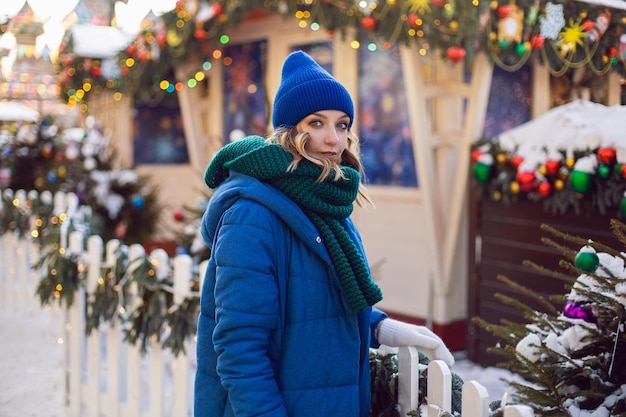 Mujer con chaqueta azul, bufanda verde y guantes blancos en invierno, en una plaza de Moscú el día de Navidad, junto a una valla blanca