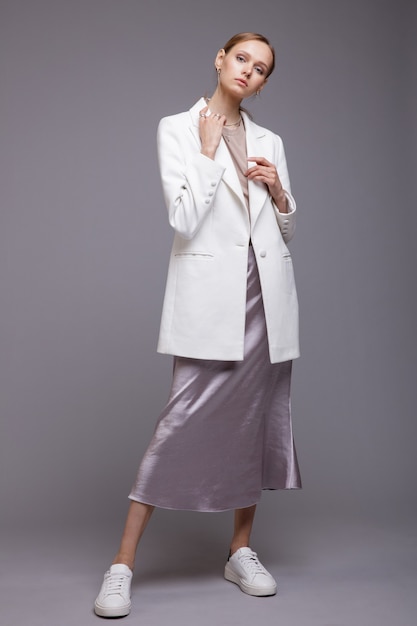 Mujer con chaqueta blanca, falda larga plateada metálica, zapatillas de deporte, fondo gris, retrato de disparo de estudio