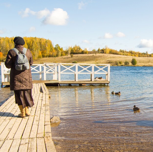 Una mujer con chaqueta y botas, con mochila, camina por un muelle de madera en un parque natural y admira las aves. Patos en el agua alrededor.