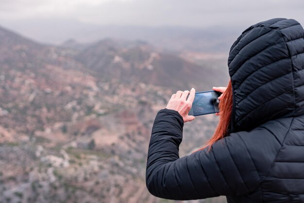 Mujer con chaqueta y capucha toma una foto del valle con su teléfono móvil Fotografía