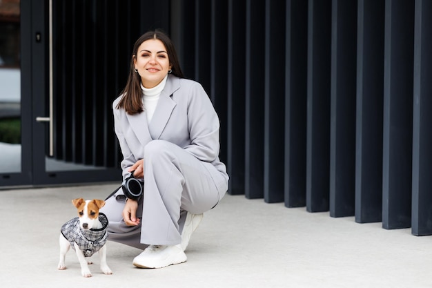 Mujer con chaqueta informal sentada cerca de su perro Jack Russell Terrier de pura raza entrenado y esponjoso favorito vestido con traje para perros cerca de un café al aire libre en el día de otoño