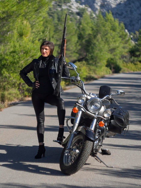 Una mujer con una chaqueta de motociclista de cuero negro con un rifle de carabina en una motocicleta chopper en Grecia