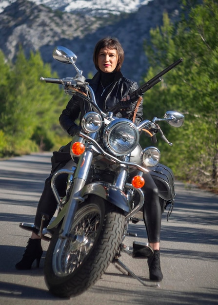 Una mujer con una chaqueta de motociclista de cuero negro con un rifle de carabina en una motocicleta chopper en Grecia