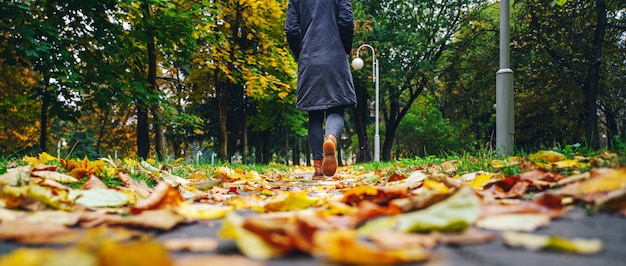 Una mujer de chaqueta negra caminando en un parque a lo largo de la acera cubierta de hojas caídas.
