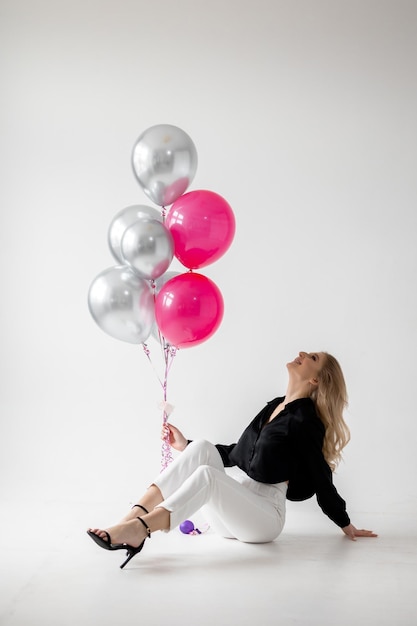 Una mujer con una chaqueta negra y pantalones blancos se sienta frente a un montón de globos rosas y plateados.