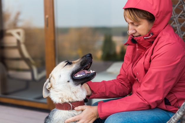 Una mujer con chaqueta roja juega y acaricia a un perro pastor mientras está sentada en una silla de mimbre