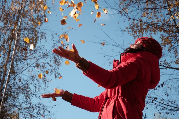 Una mujer con una chaqueta roja lanza hojas amarillas. Paisaje de otoño. Video en cámara lenta