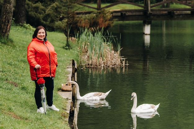 Una mujer con una chaqueta roja y un paraguas camina por un parque de verano cerca de un lago con cisnes.