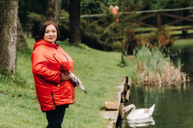 Una mujer con una chaqueta roja y un paraguas camina por un parque de verano cerca de un lago con cisnes.