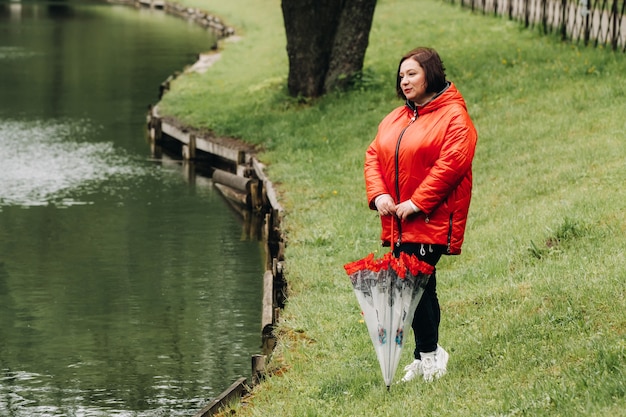 Una mujer con una chaqueta roja y con un paraguas camina en el parque de verano cerca del lago