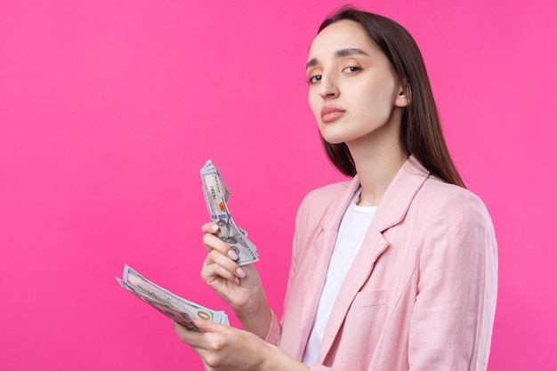 Una mujer con una chaqueta rosa tiene billetes en dólares, una chica de negocios desconcertada esconde dólares sobre un fondo rojo.