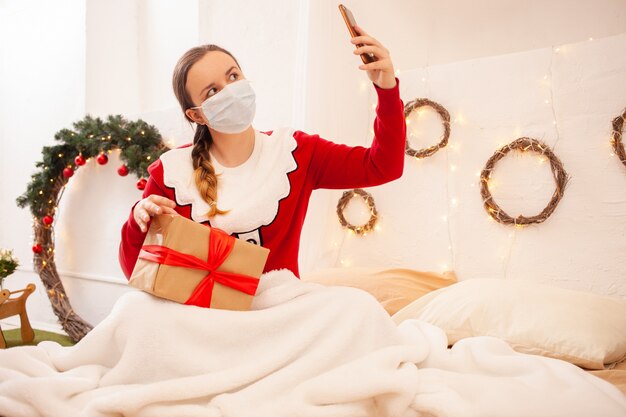 Mujer en chaqueta de santa claus sostiene teléfono y se comunica con sus amigos en máscaras protectoras. Celebración remota de año nuevo y navidad