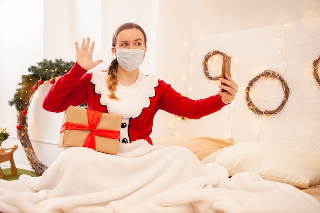 Mujer en chaqueta de santa claus sostiene teléfono y se comunica con sus amigos en máscaras protectoras. Celebración remota de año nuevo y navidad